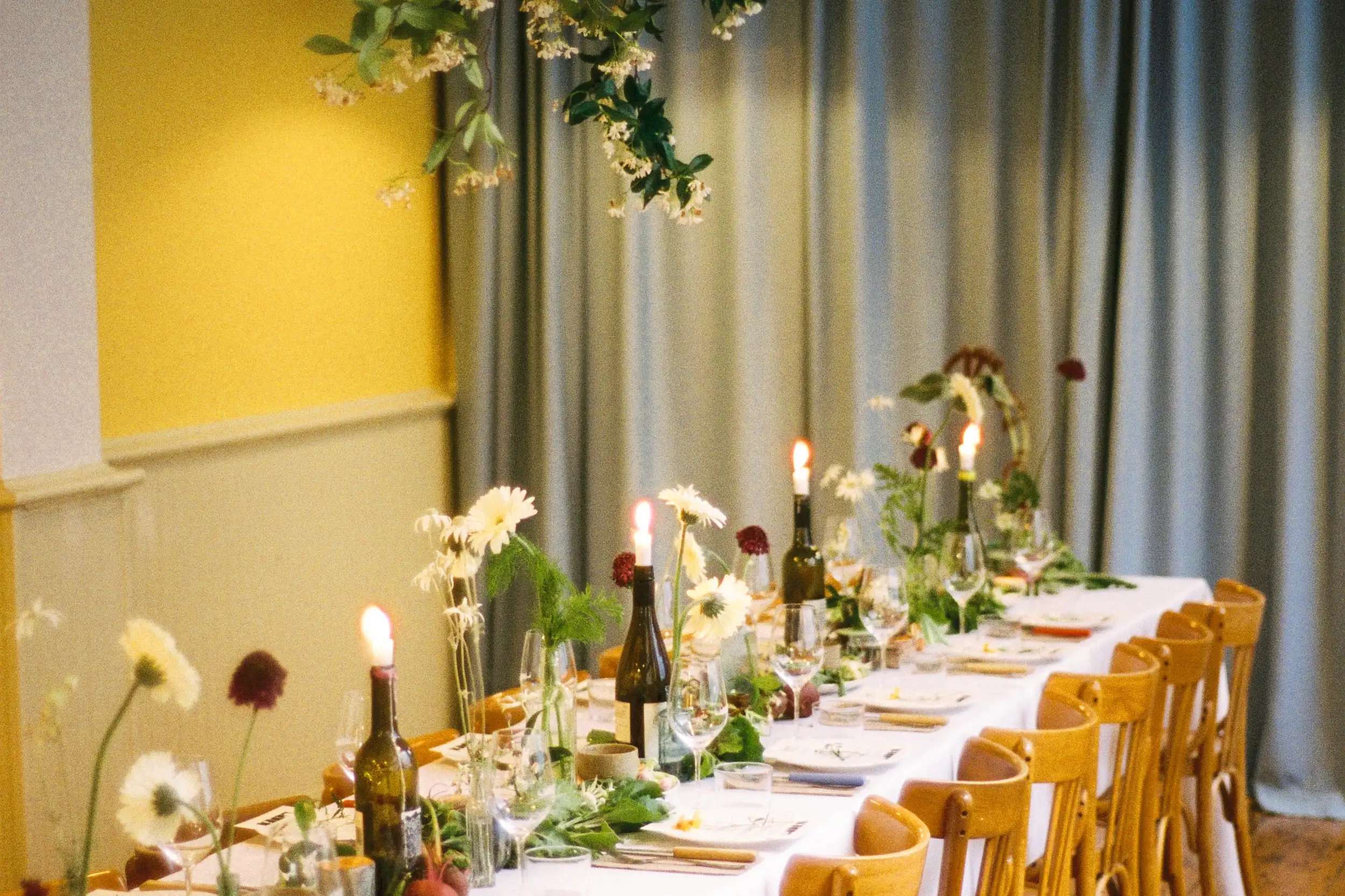 Long dining table set with white tablecloth, wine bottles with lit candles, glassware, and floral decorations under hanging greenery.