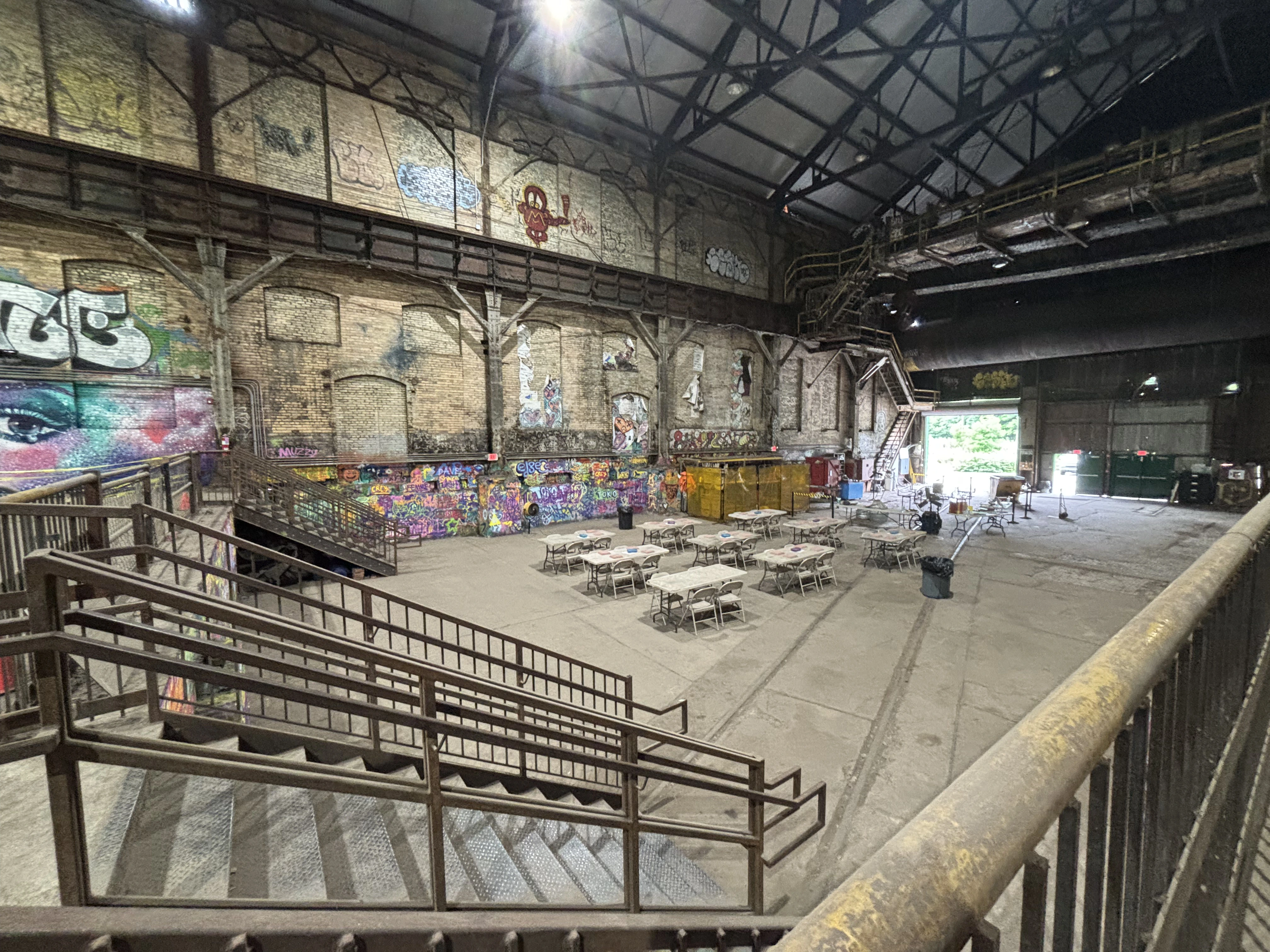 Interior of a large industrial building with graffiti-covered brick walls and rows of folding tables and chairs arranged on the concrete floor.