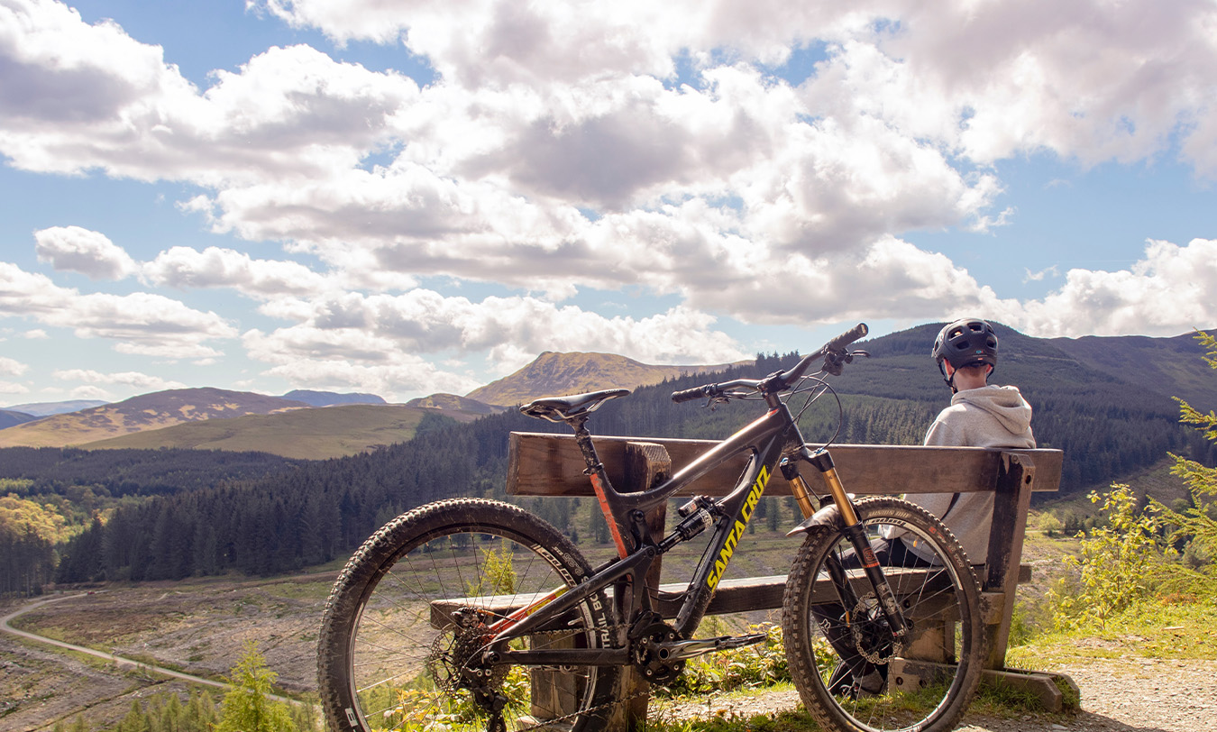 A man sat on a bench looking at a view of the Lake District. A bike is leaning against the bench.