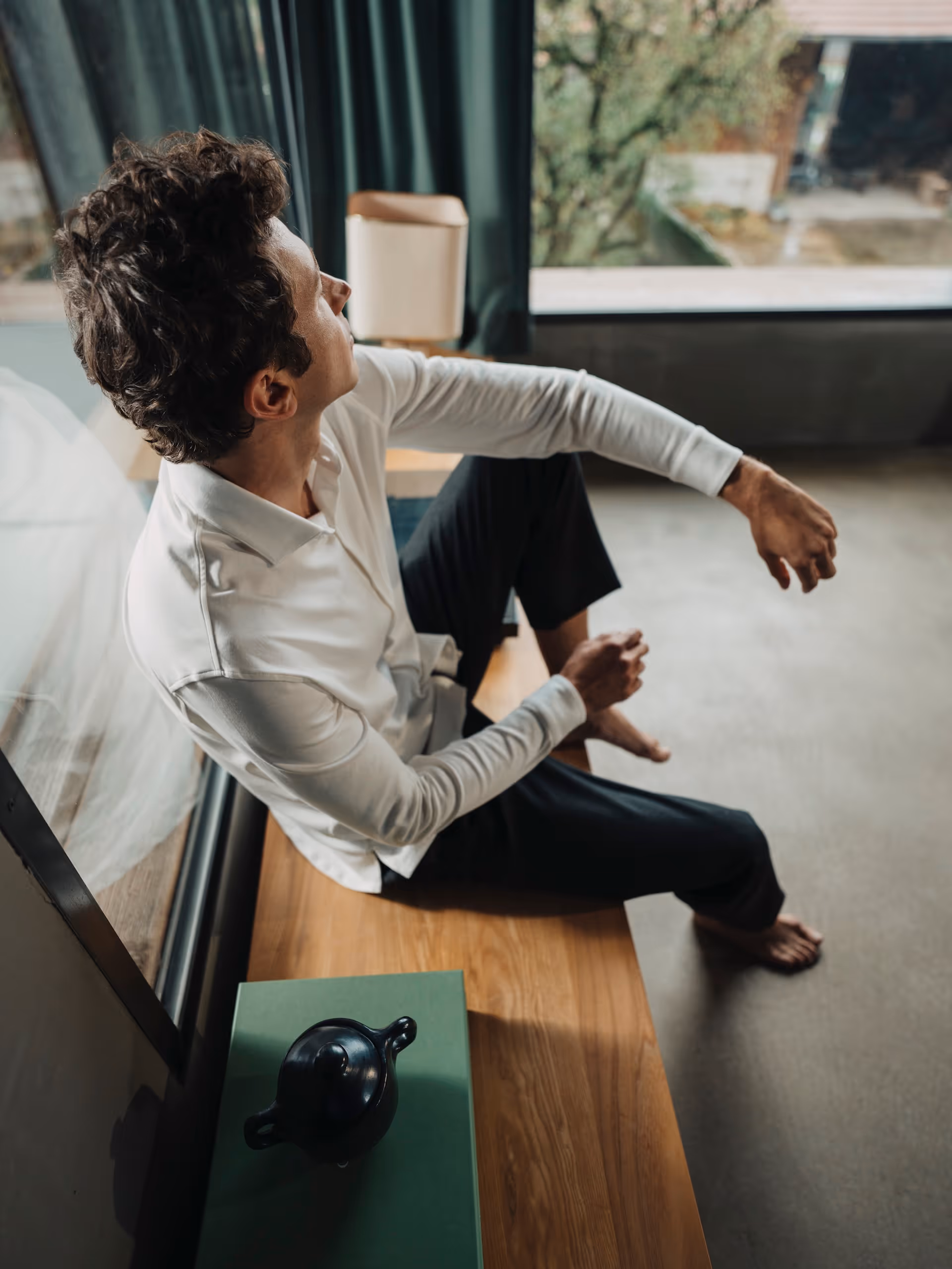 Man sitting by a window in a modern room, wearing a white Chirimoya Pima cotton dress shirt and leisure trousers.