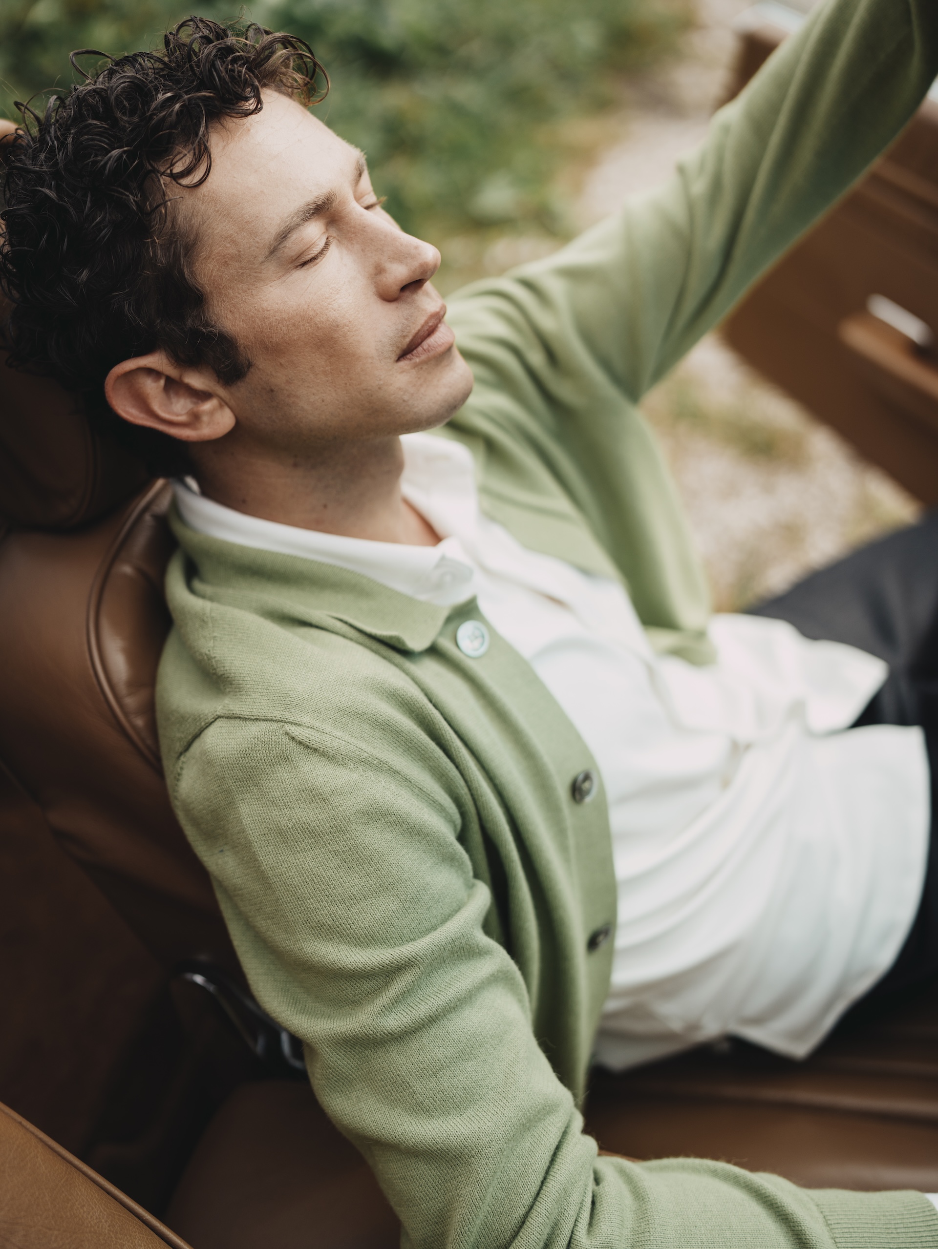 Man relaxing with eyes closed in a vintage car, wearing a green Chirimoya Baby Pima cardigan and white Pima shirt.