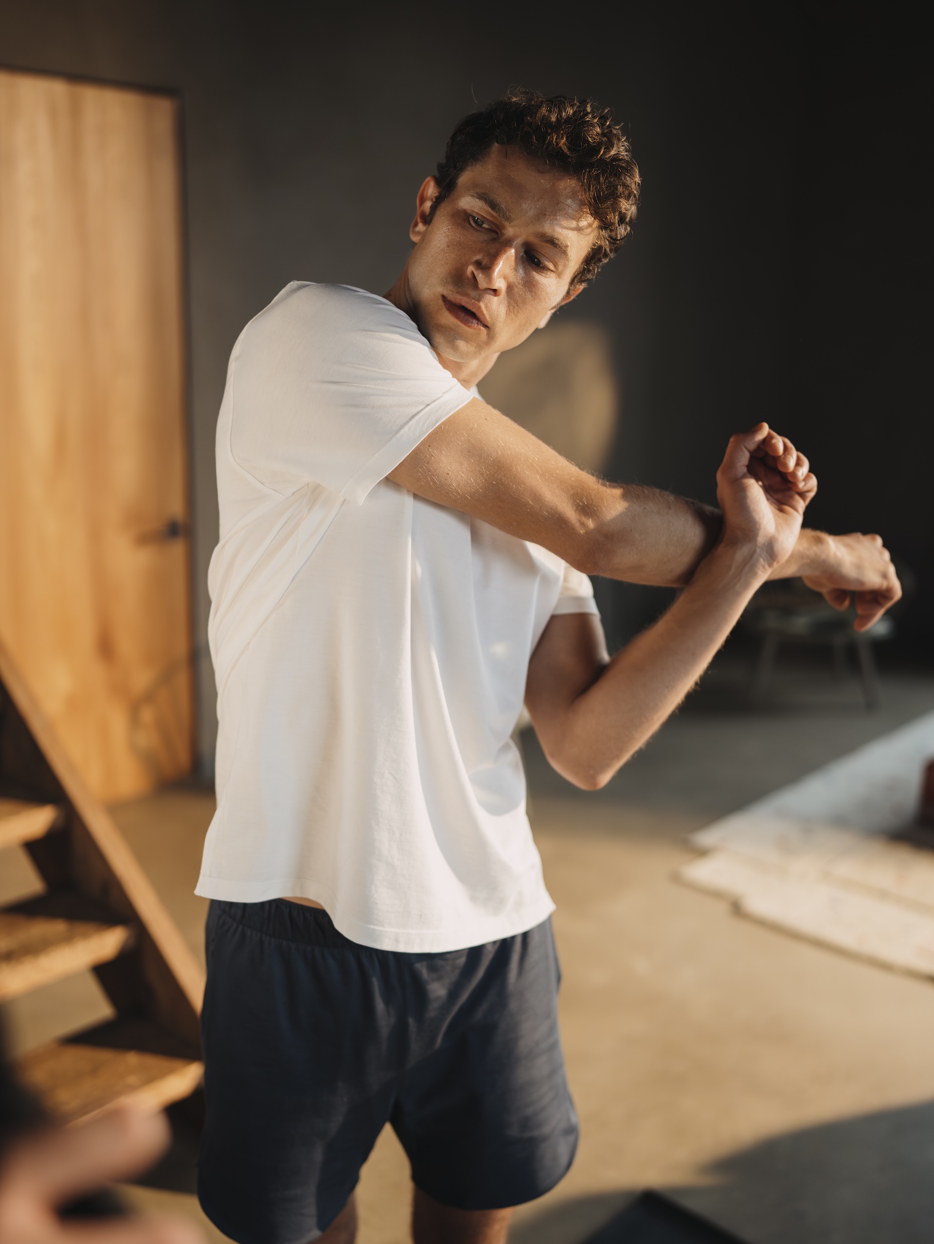 Man stretching indoors wearing a white Chirimoya Classic Pima cotton T-shirt and navy yoga shorts.
