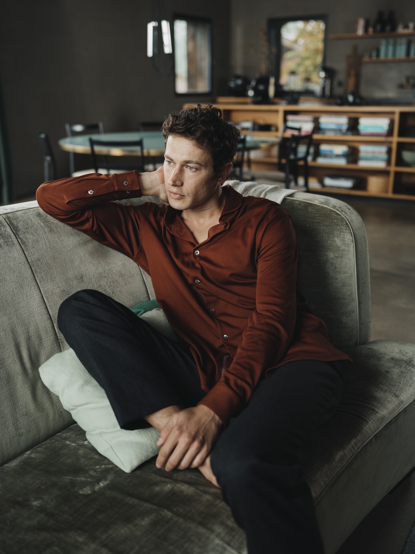 Man sitting on a couch wearing a mahogany Chirimoya Natural Dye Pima dress shirt and black yoga trousers.