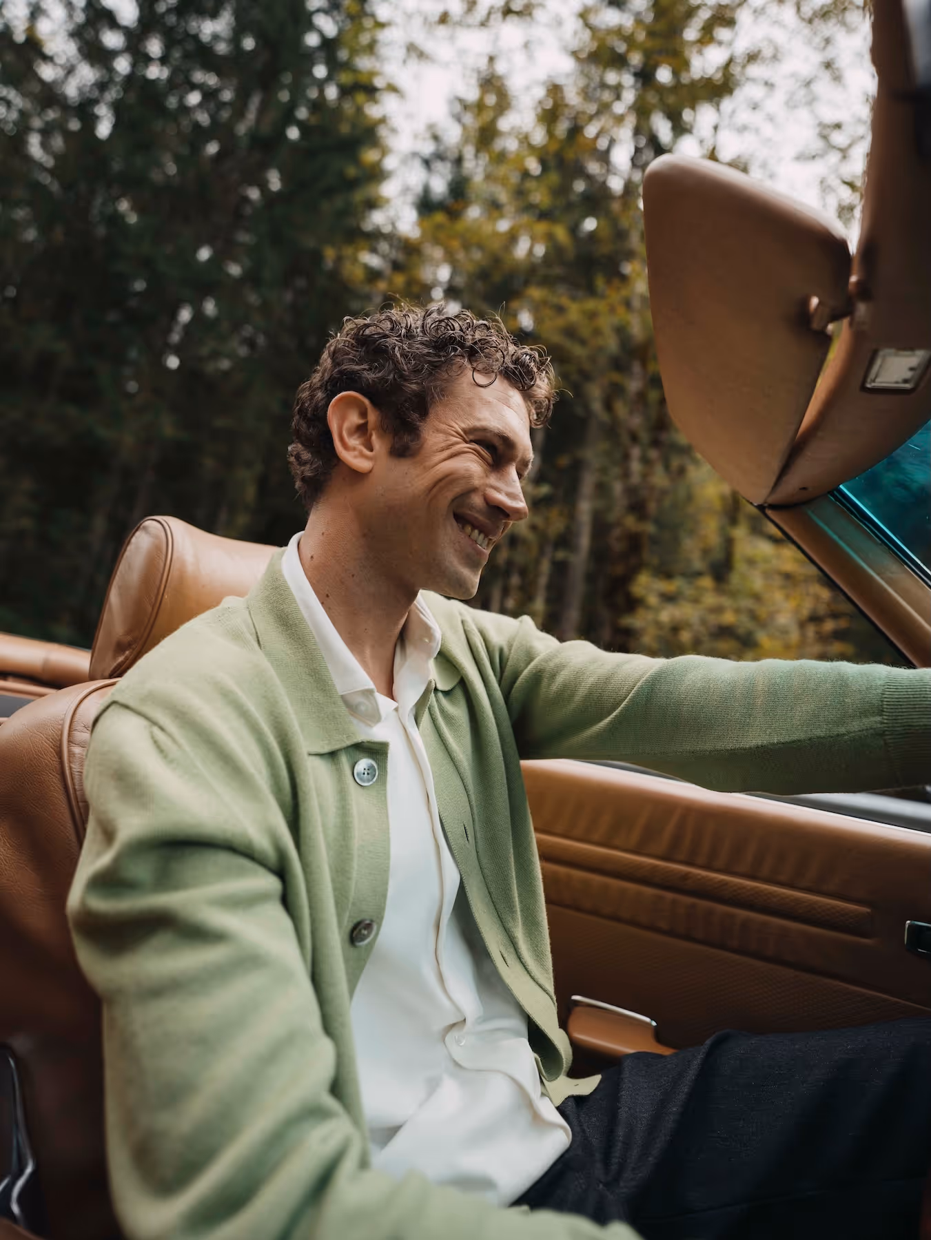 Man driving a vintage car, wearing a green Chirimoya Baby Pima cardigan and white Pima shirt.