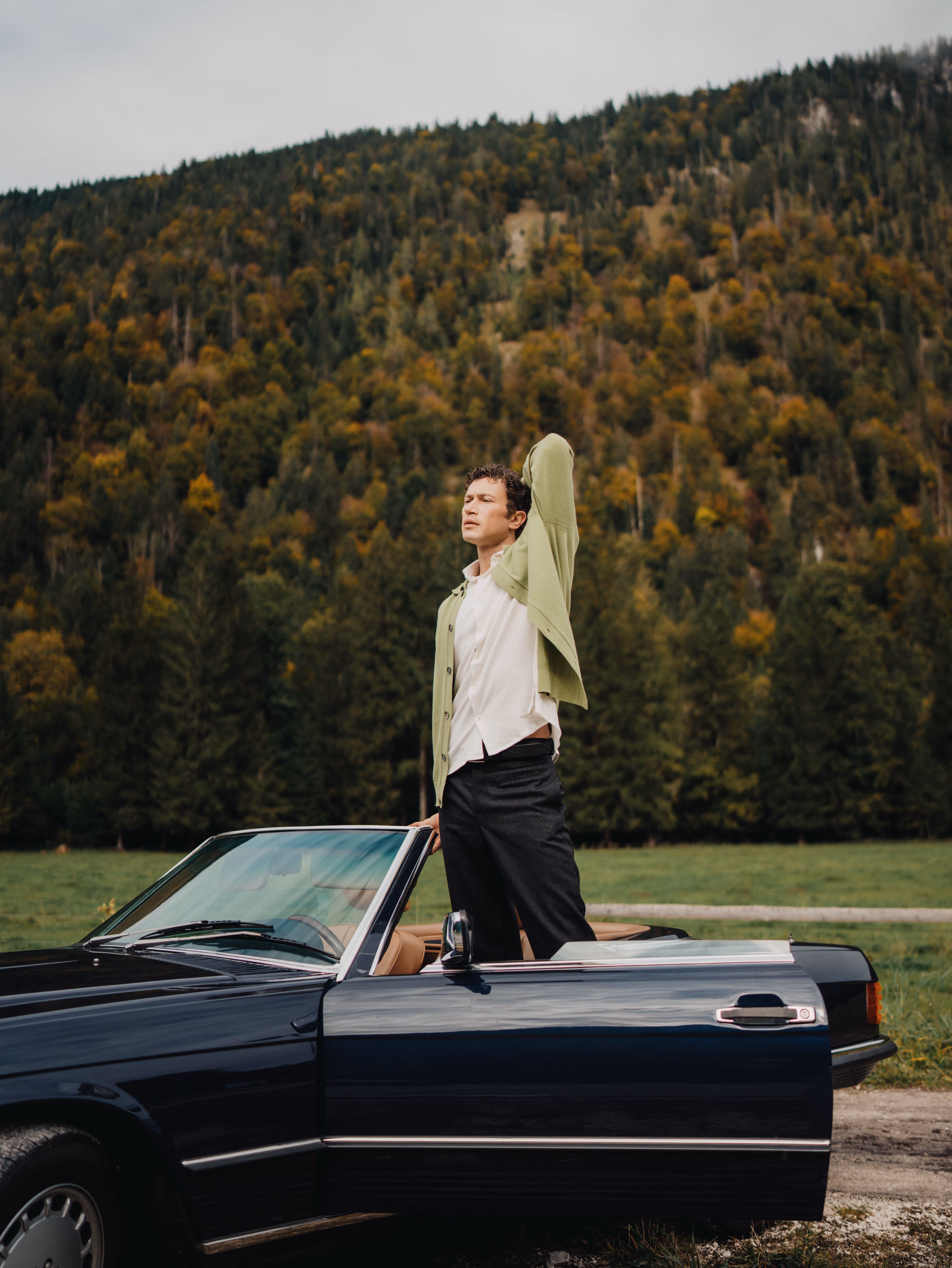 Man wearing a Chirimoya baby pima cardigan and pima dress shirt, standing beside a vintage car in a mountain landscape.