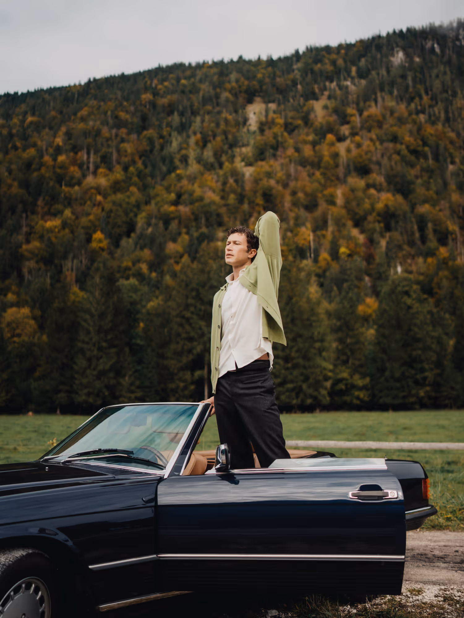 Man wearing a Chirimoya baby pima cardigan and pima dress shirt, standing beside a vintage car in a mountain landscape.