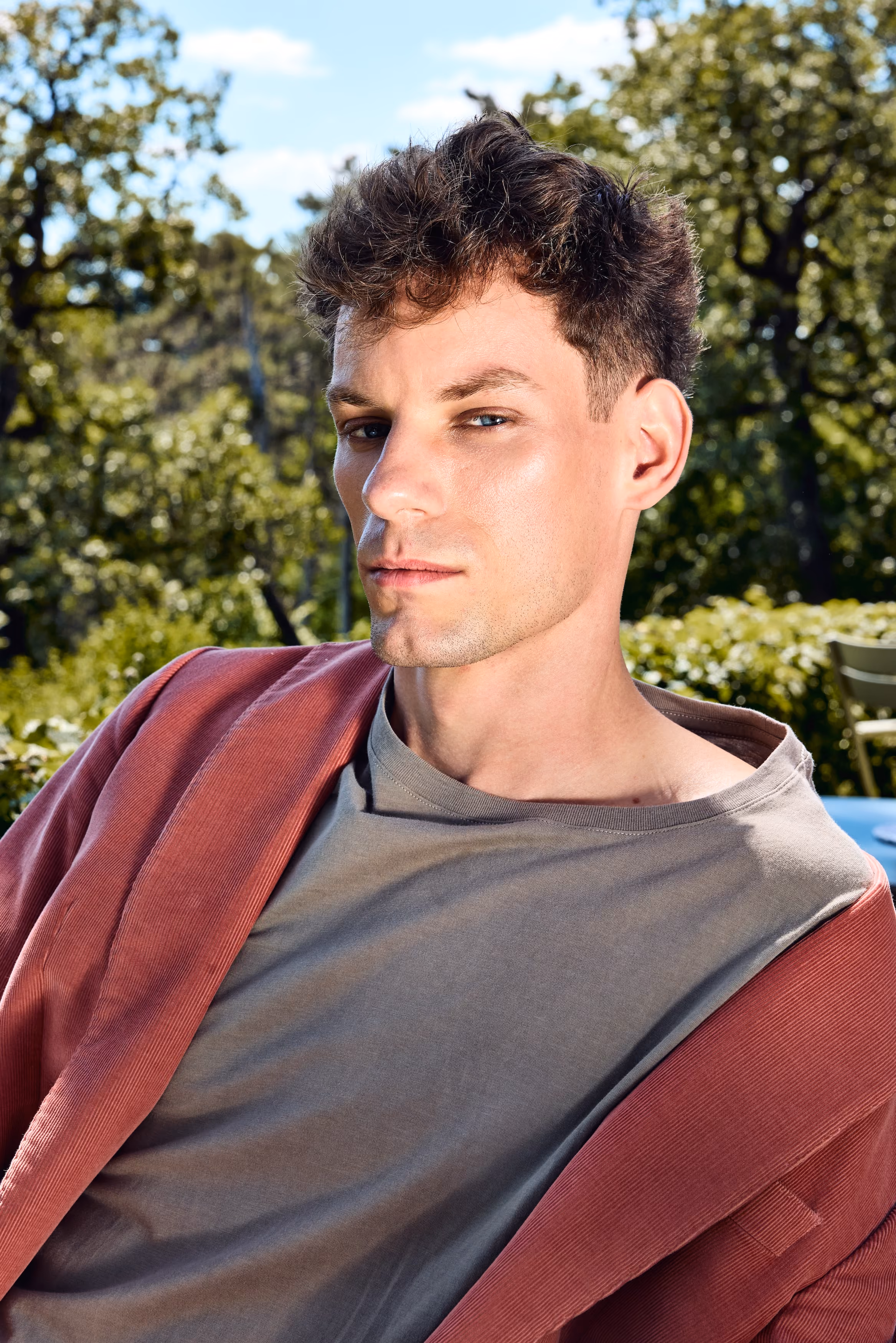 Man with short curly hair wearing a gray shirt and red jacket outdoors with trees behind him.
