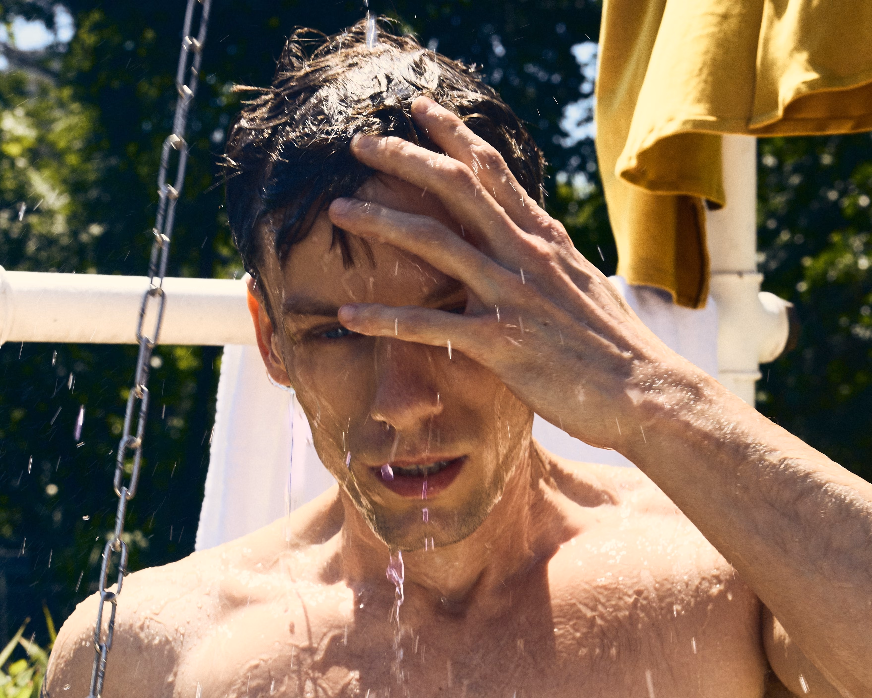 Shirtless man rinsing his wet hair outdoors under a shower with water dripping down his face.