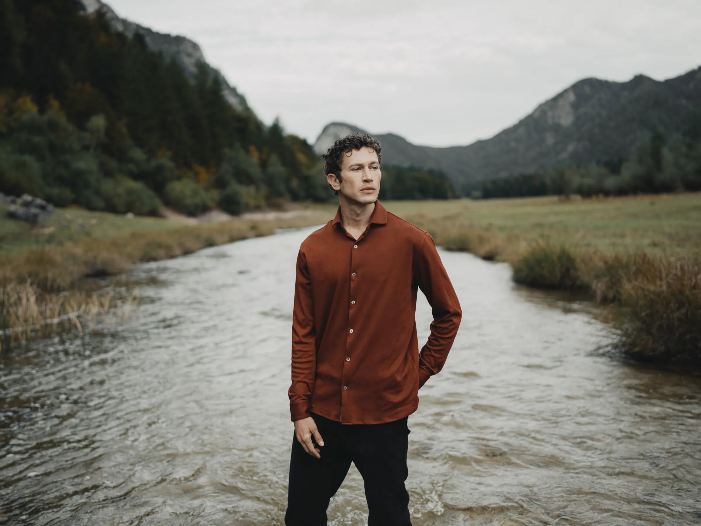 Man in a rust-coloured shirt standing in a shallow mountain stream