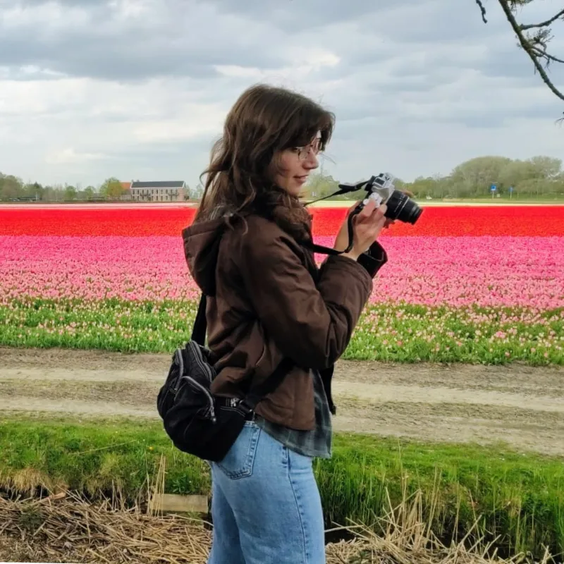 A young woman stands on a rocky forest path, holding a camera up to their face while photographing the surrounding landscape.