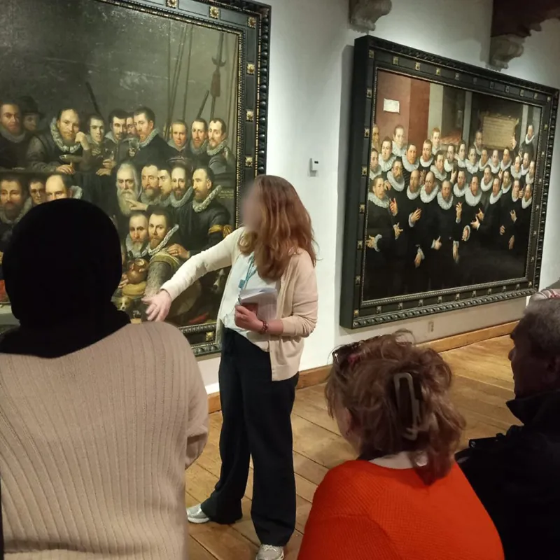 A group of people viewing large, classical oil portraits in an art gallery. A museum guide with long hair and a light cardigan stands in the center, gesturing toward one of the paintings while holding a guidebook.