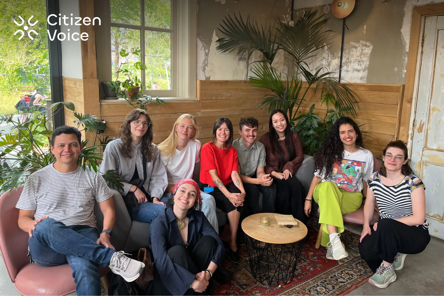 The Citizen Voice team, a diverse group of 9 people, relaxing together on couches in a cozy, plant-filled café space.