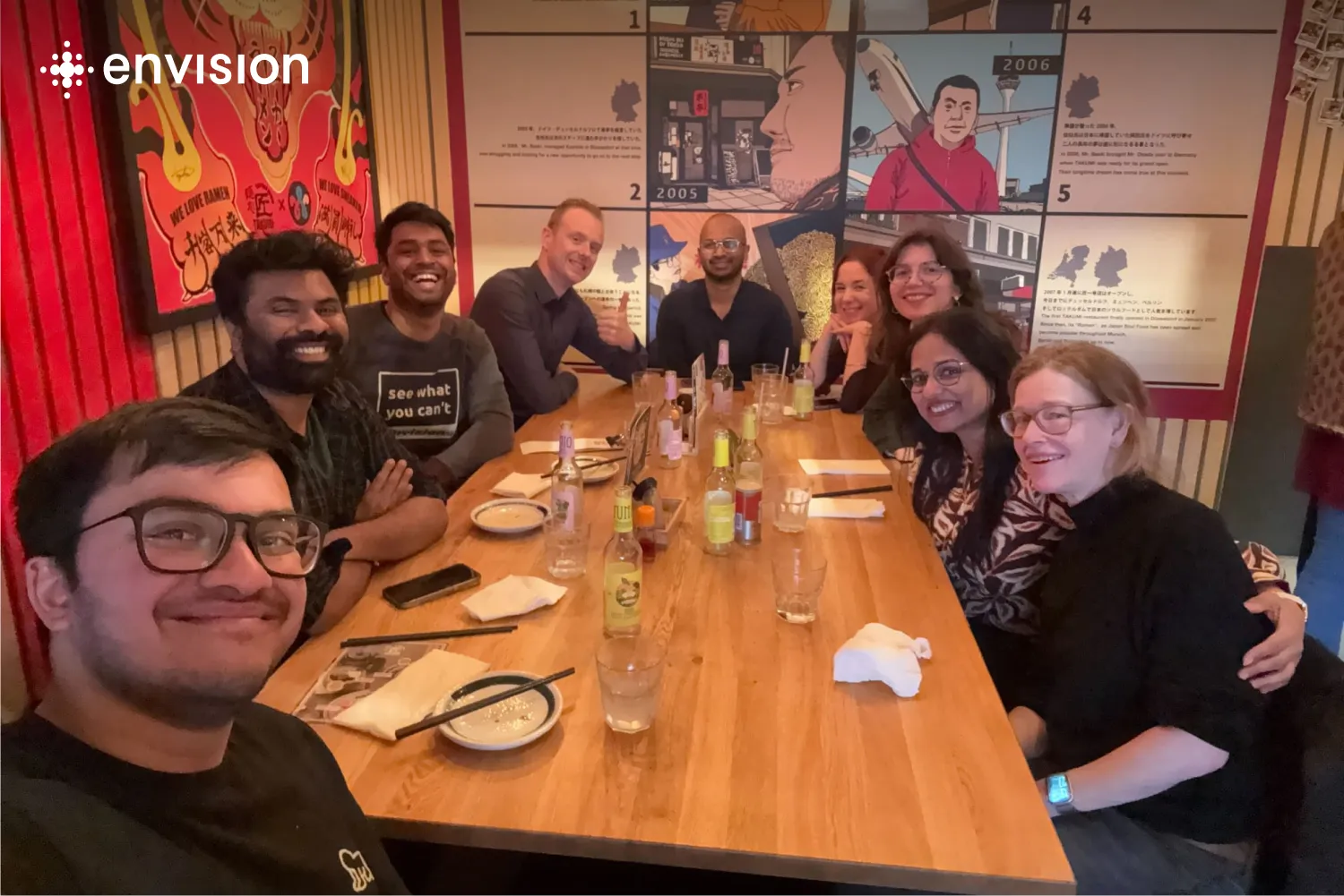 The Envision team, a diverse group of 9 people, gathered around a restaurant table at a ramen spot, smiling for a group photo.