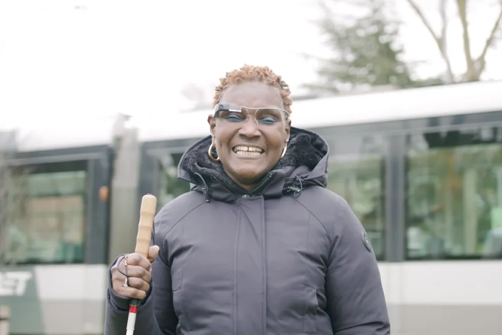 A smiling woman wearing smart glasses and a dark puffer jacket holds a white cane while standing outdoors in front of a bus or tram, on an overcast day.