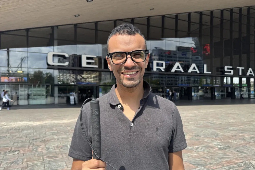 A smiling man wearing black-framed smart glasses and a gray polo shirt stands in front of Rotterdam Centraal train station on a sunny day, holding a cane in hist right hand.