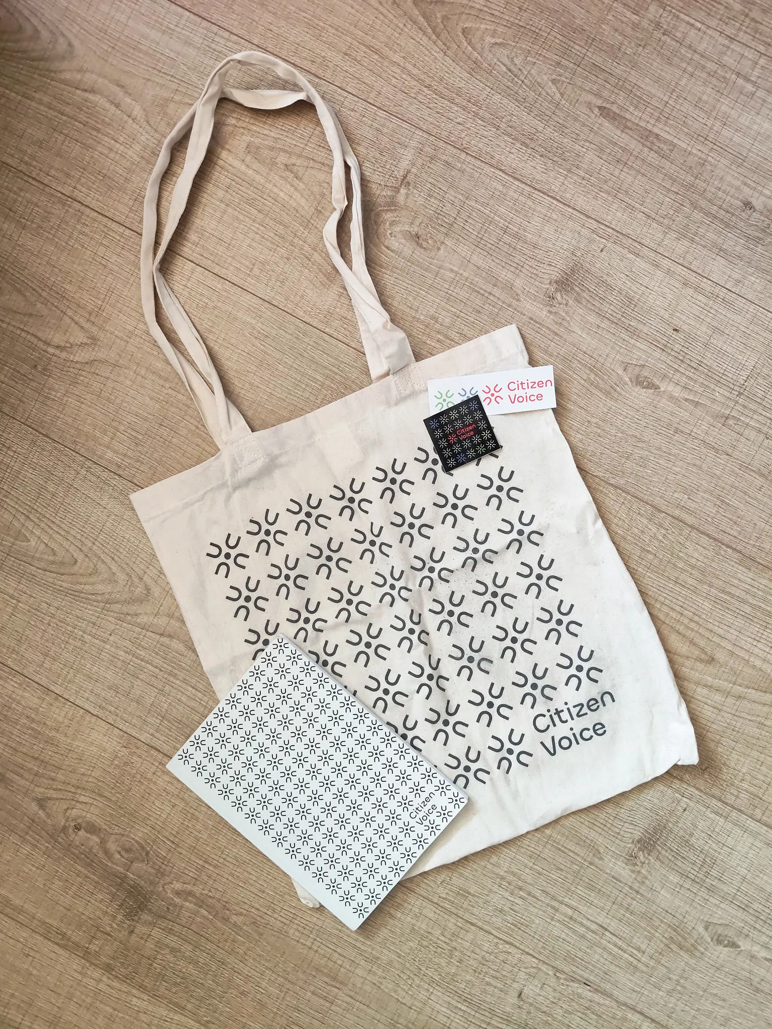 A cream-colored canvas tote bag printed with a repeating pattern of the Citizen Voice logo mark and the words "Citizen Voice," laid flat on a wooden floor alongside a matching notebook and a set of stickers following the same motif.