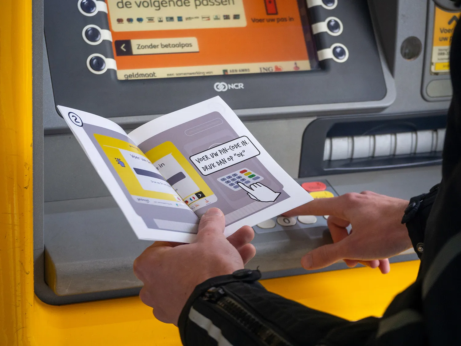 A person's hands hold open a small illustrated instruction booklet in front of a yellow Geldmaat ATM machine. The open page shows a step-by-step diagram with text reading "Voer uw PIN-code in, druk dan op OK" alongside an illustration of a hand pressing a keypad.