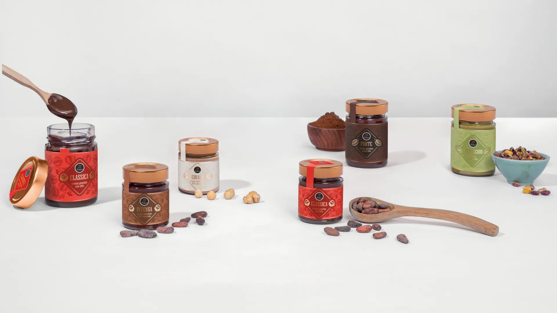 A product photography shot of six Bodrato gianduja spread jars arranged on a white surface, with copper lids and varied label colors — red, cream, dark brown, and green — labeled Classica, Intensa, Chiara, Forte, and Pistacchio. Scattered around them are cacao beans, hazelnuts, ground cacao powder, and pistachios, with a wooden spoon dripping chocolate spread in the foreground.