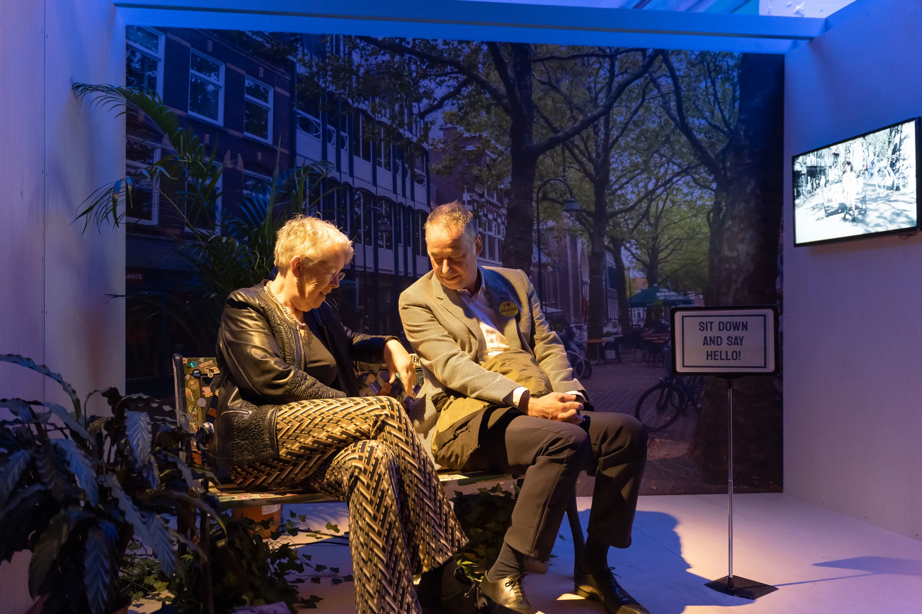 Two visitors — a woman and a man — sit on a decorative bench inside a dimly lit installation space. Behind them is a large-scale mural photograph of a Dutch canal street. A sign to their right reads "Sit down and say hello!" and a monitor plays video in the background.