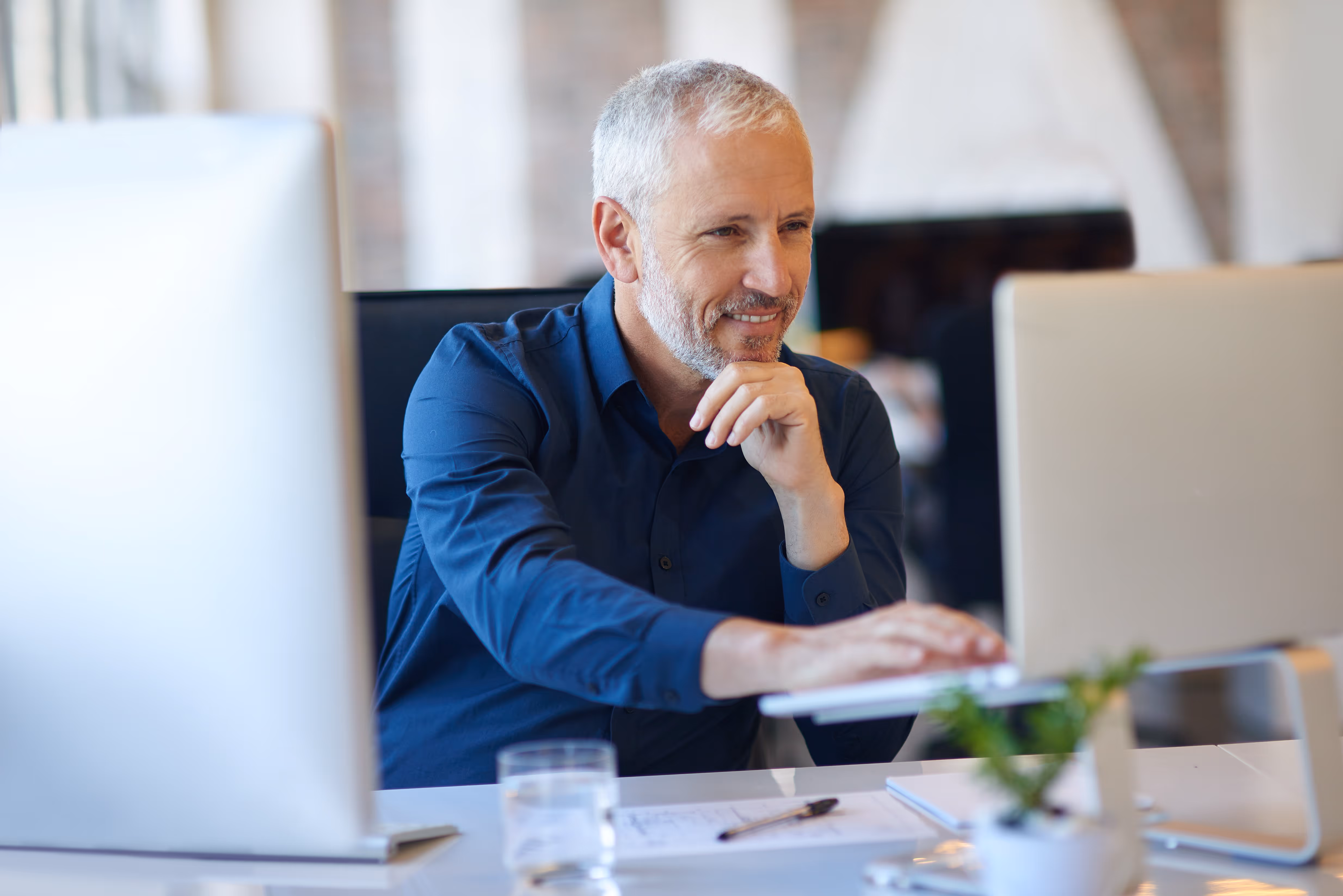 En glad forretningsmand arbejder ved sit skrivebord med flere computere og skærme i et moderne kontormiljø.