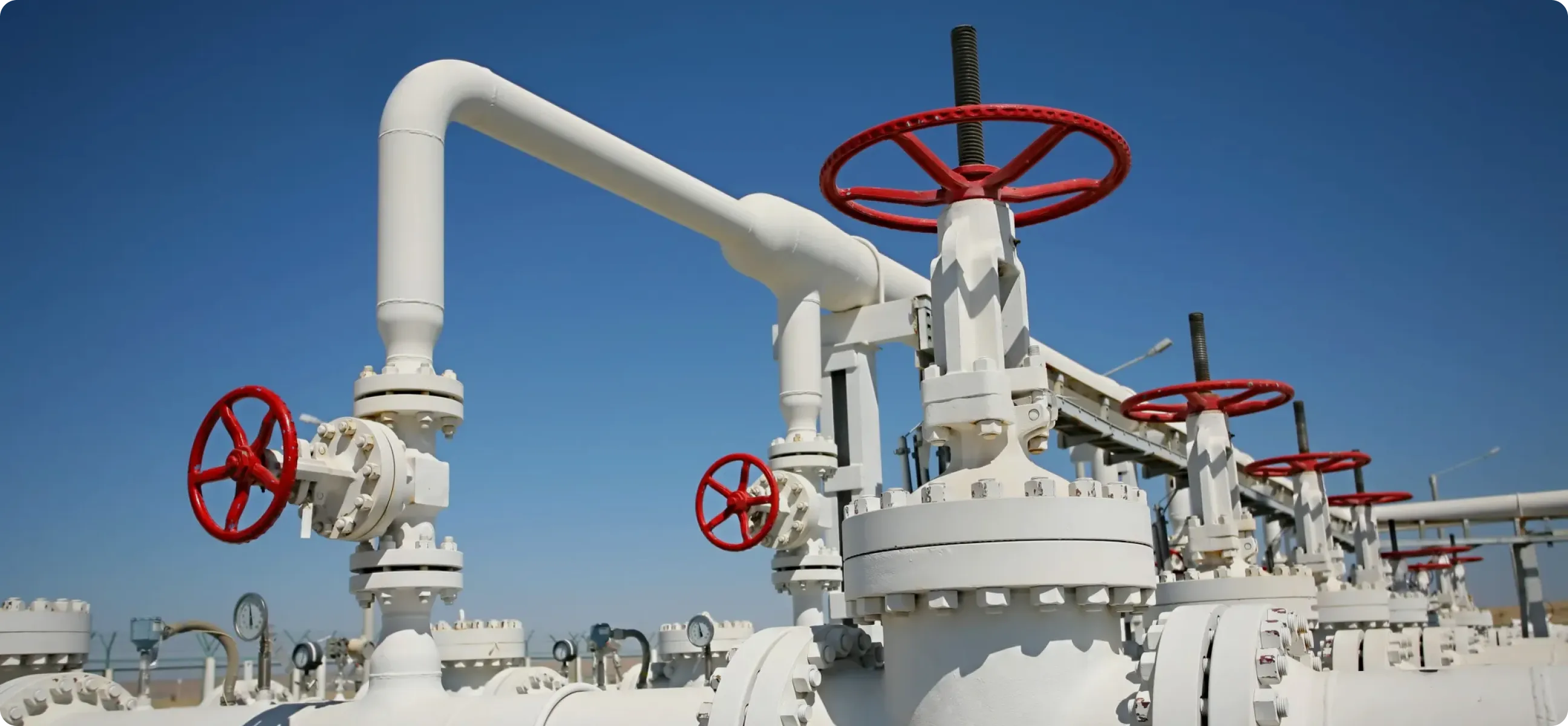 A row of large industrial white pipelines with red valve wheels and pressure gauges, set against a clear blue sky in an outdoor facility.