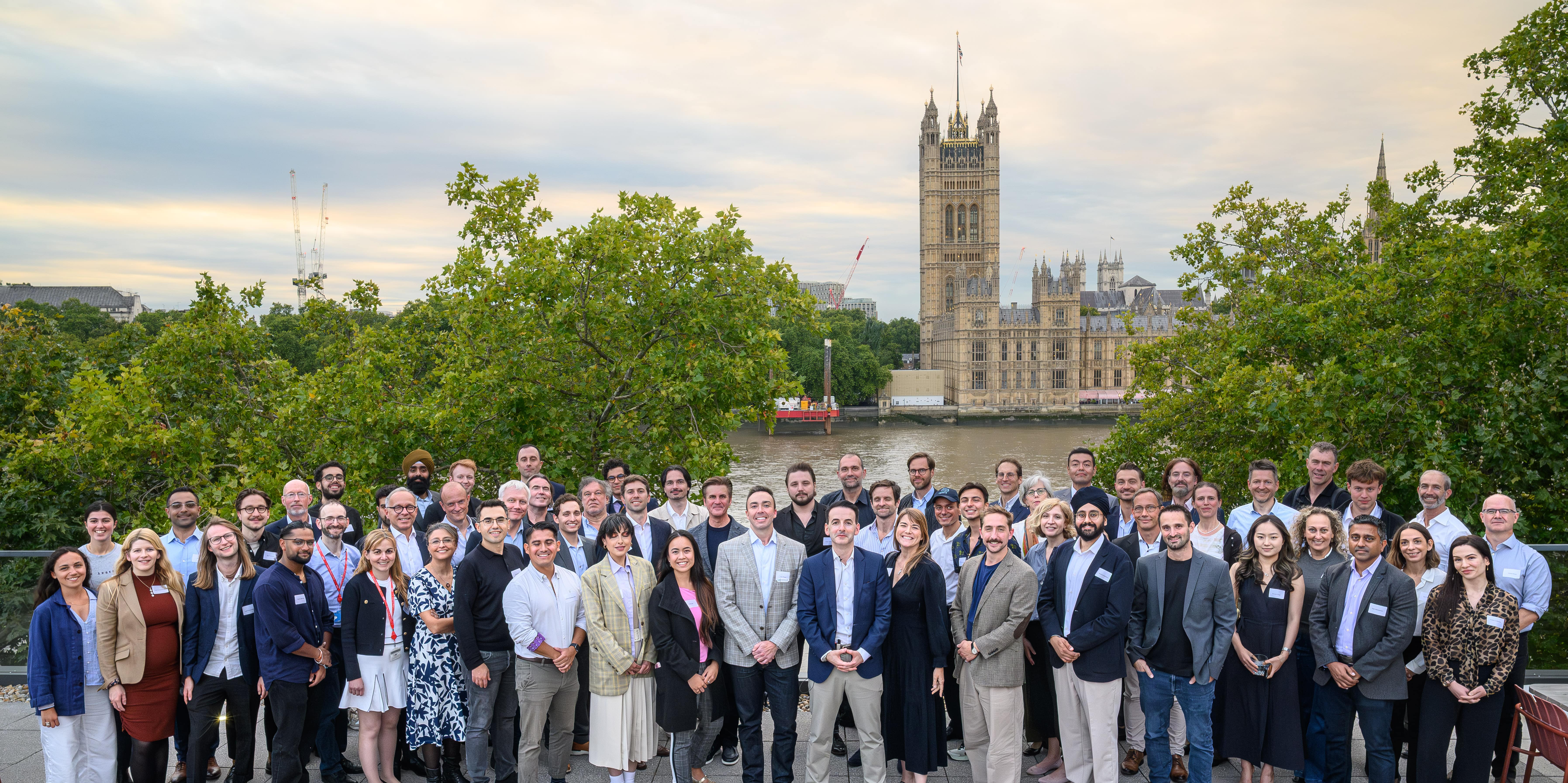 Group photo of 80 people standing on the other side of the river of Buckimgham Palace, next to the Thames River.