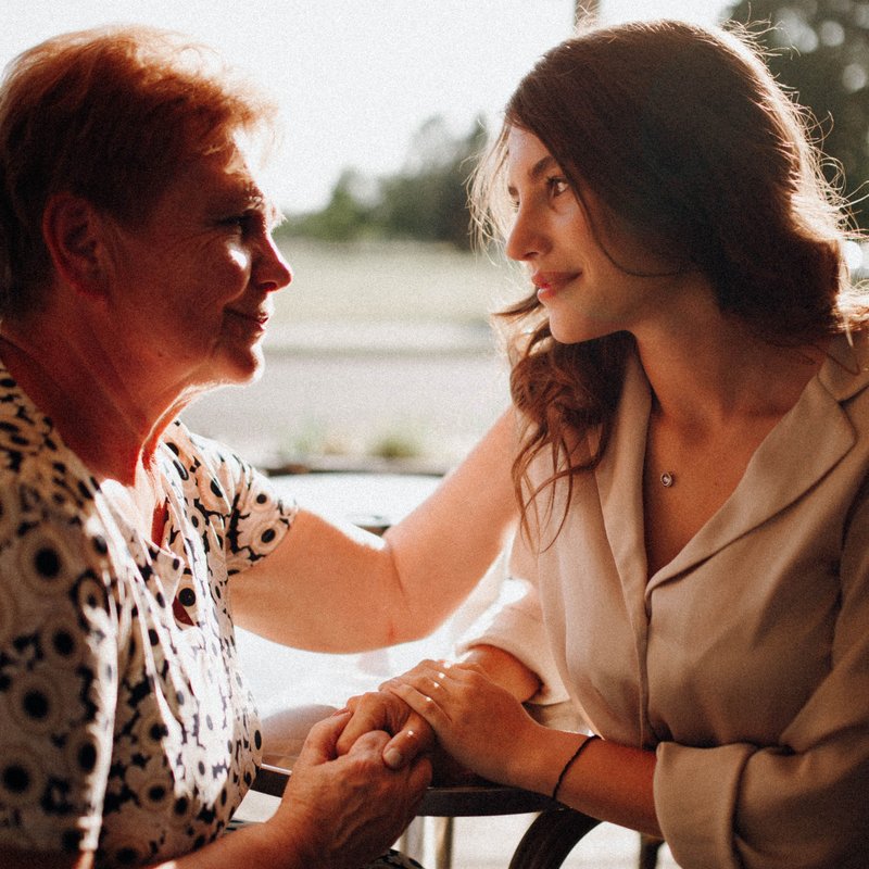 Eine Tochter und ihre Mutter sitzen einander zugewandt im Gespräch und halten sich an den Händen.