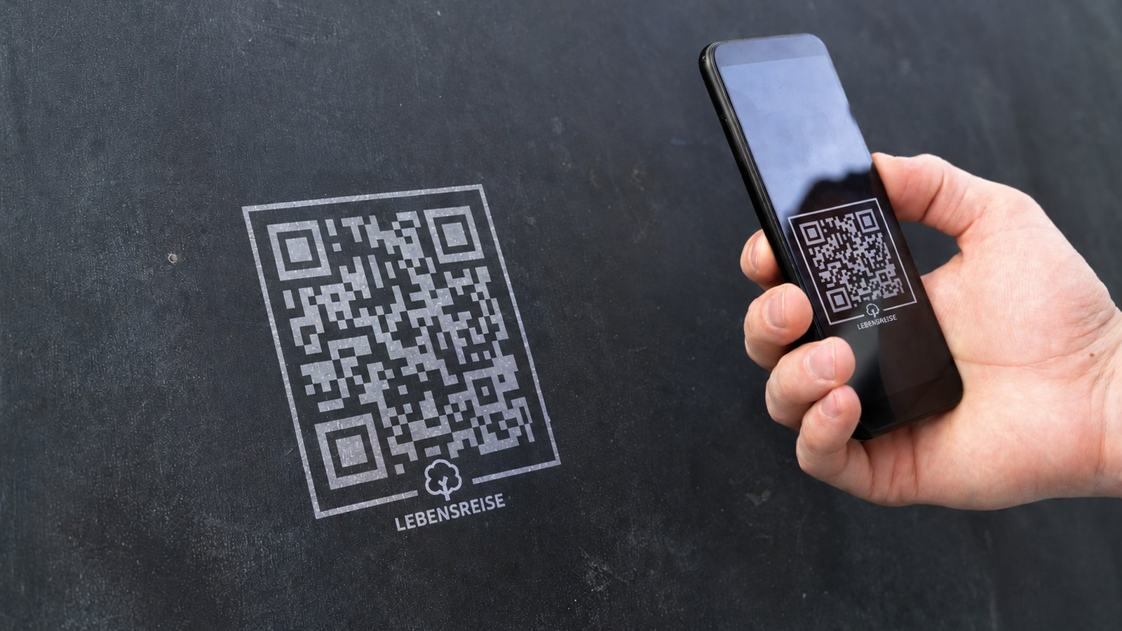 Ein QR-Code mit Lebensreise-Logo auf einer dunklen Wand, davor eine Hand mit einem Mobiltelefon, das den Code erfasst.