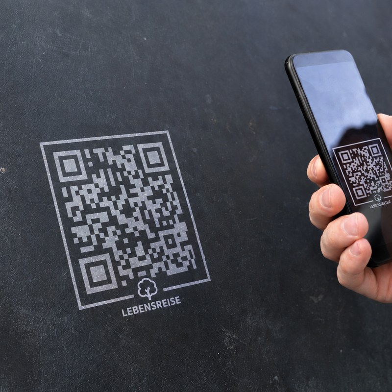 Ein QR-Code mit Lebensreise-Logo auf einer dunklen Wand, davor eine Hand mit einem Mobiltelefon, das den Code erfasst.
