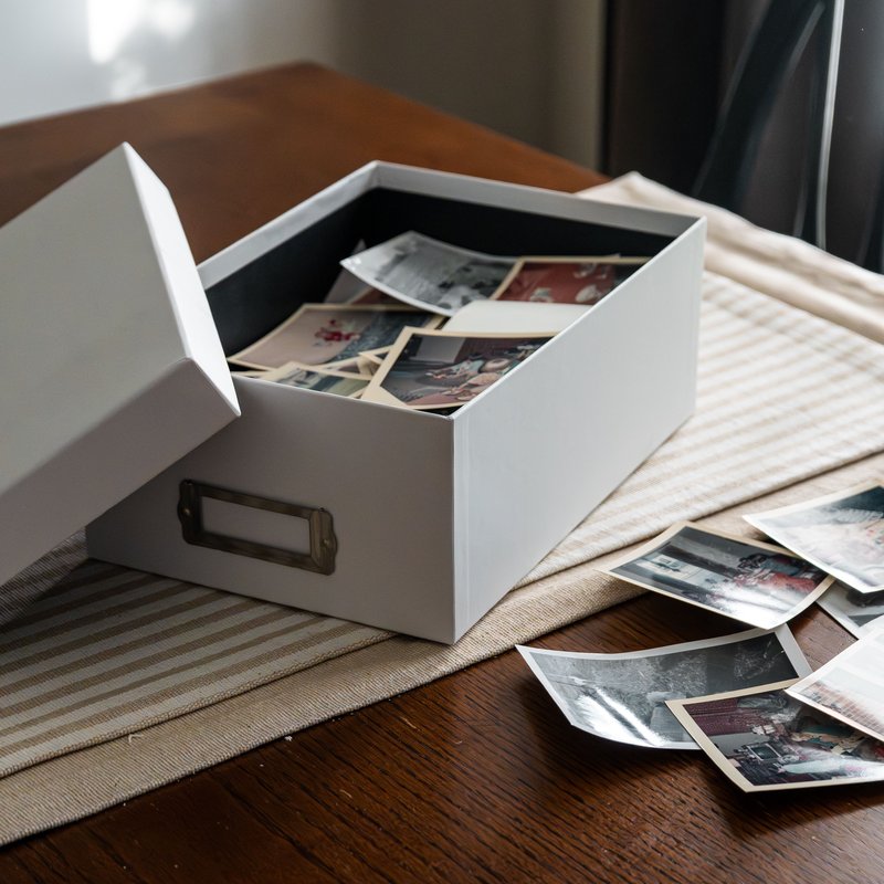 Eine geöffnete Fotoschachtel steht auf einem Holztisch, daneben liegen alte Papierfotos ausgebreitet.