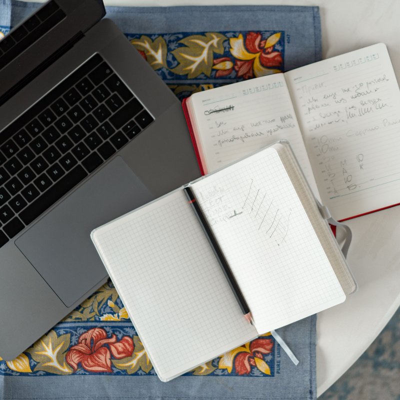 Ein Laptop und zwei aufgeschlagene Notizbücher mit Stift liegen auf einem runden Tisch.