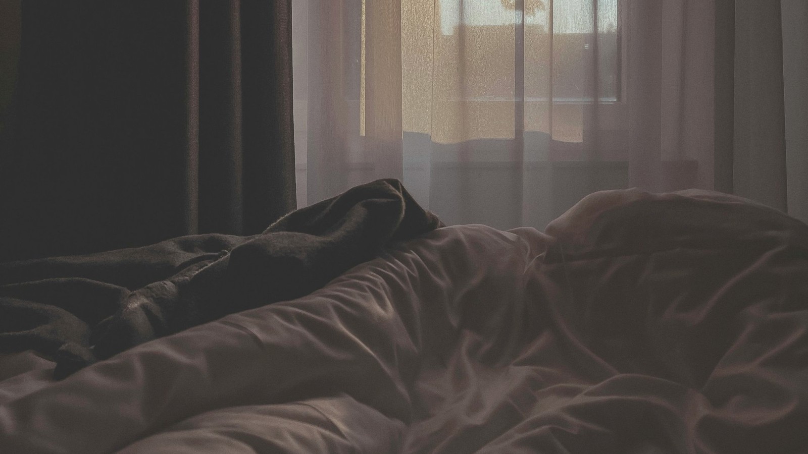 Ein zerwühltes Bett in einem abgedunkelten Zimmer, durch den Vorhang fällt Morgenlicht.