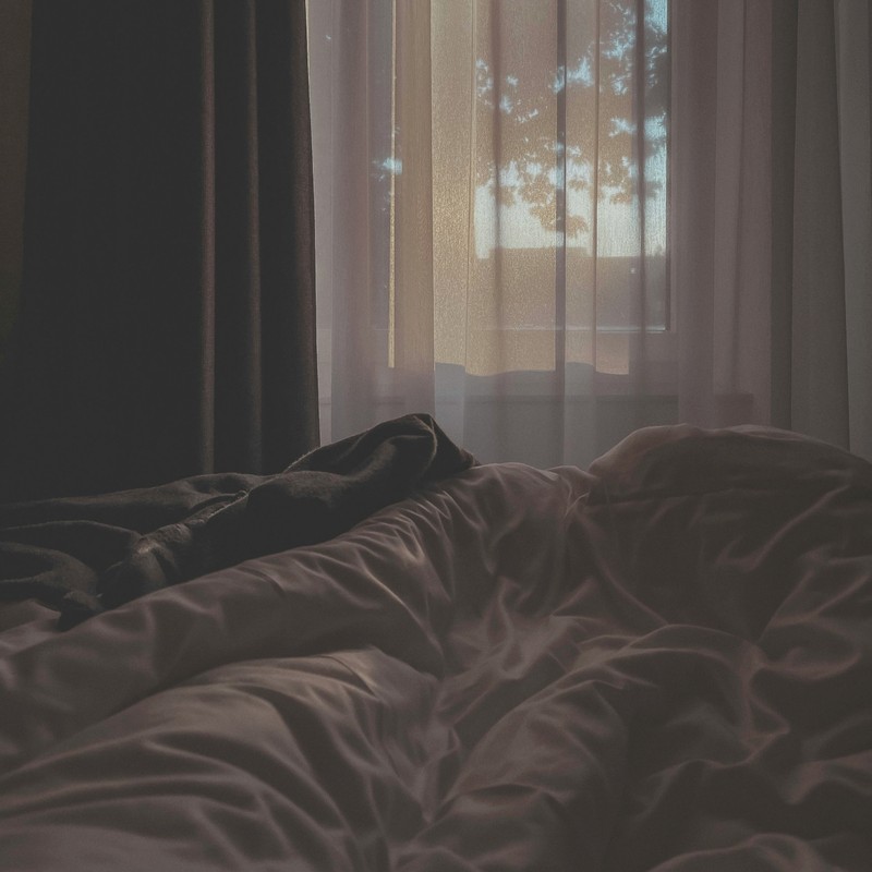 Ein zerwühltes Bett in einem abgedunkelten Zimmer, durch den Vorhang fällt Morgenlicht.