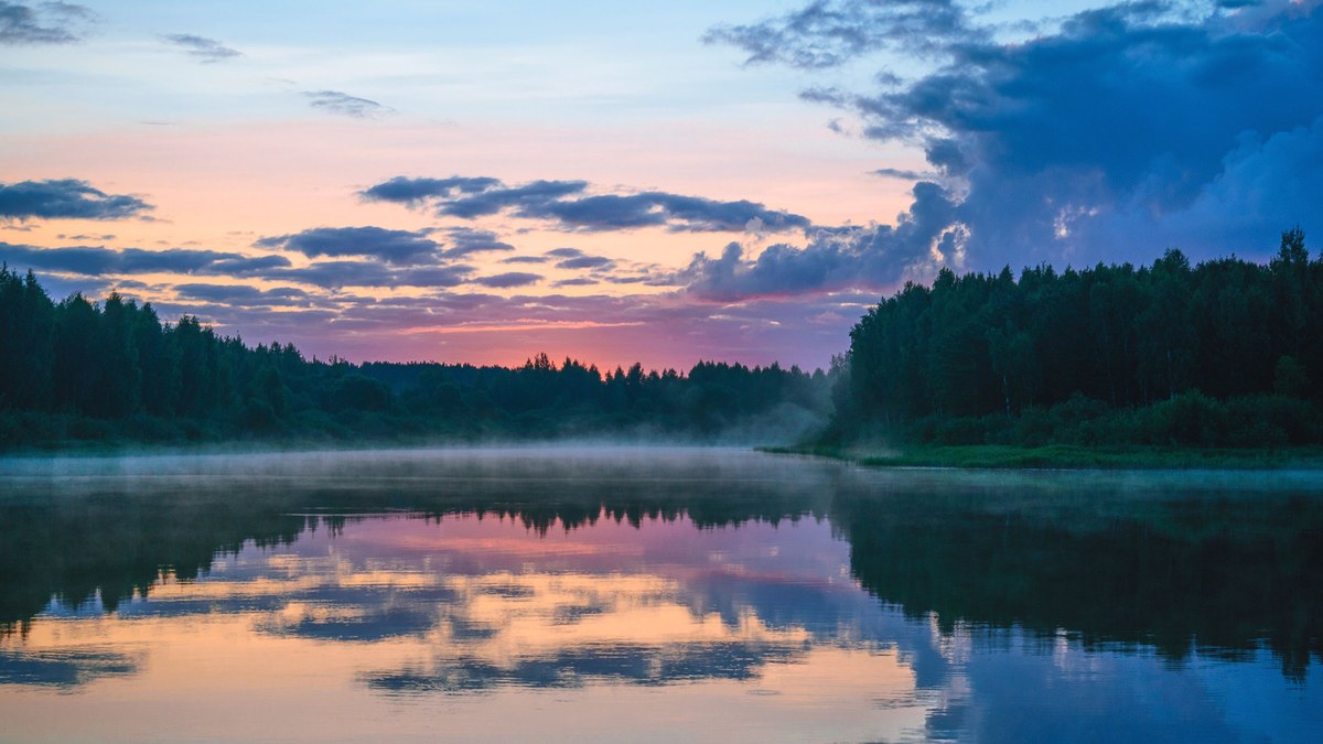 Ein ruhiger Fluss im Morgennebel, am Ufer Wald, der Himmel spiegelt sich im Wasser.