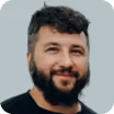 Smiling man with dark hair and beard wearing a black shirt on light gray background.