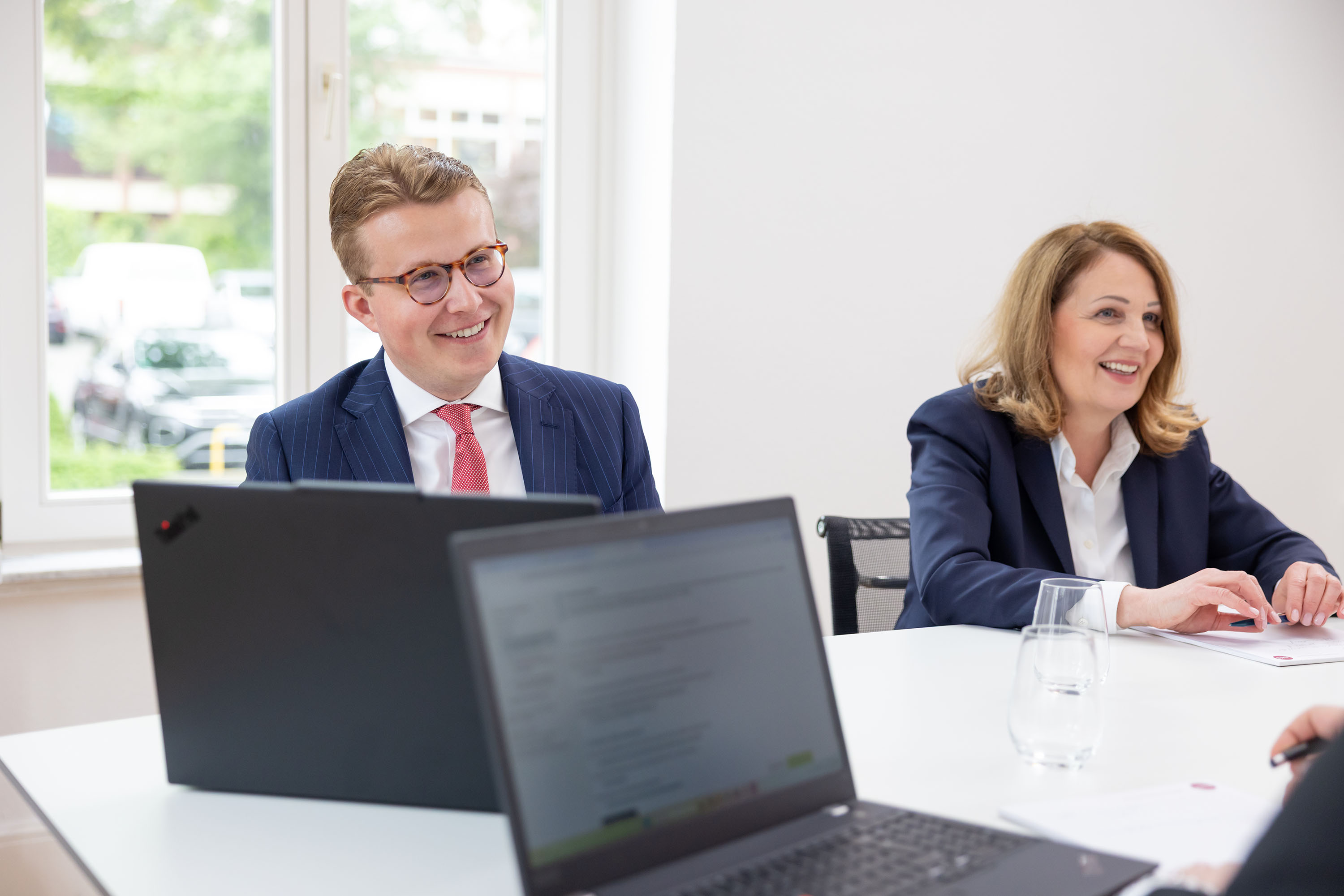 Mann im Nadelstreifenanzug und Frau im Blazer sitzen nebeneinander, lächeln und schauen in Richtung weiterer Gesprächspartner.