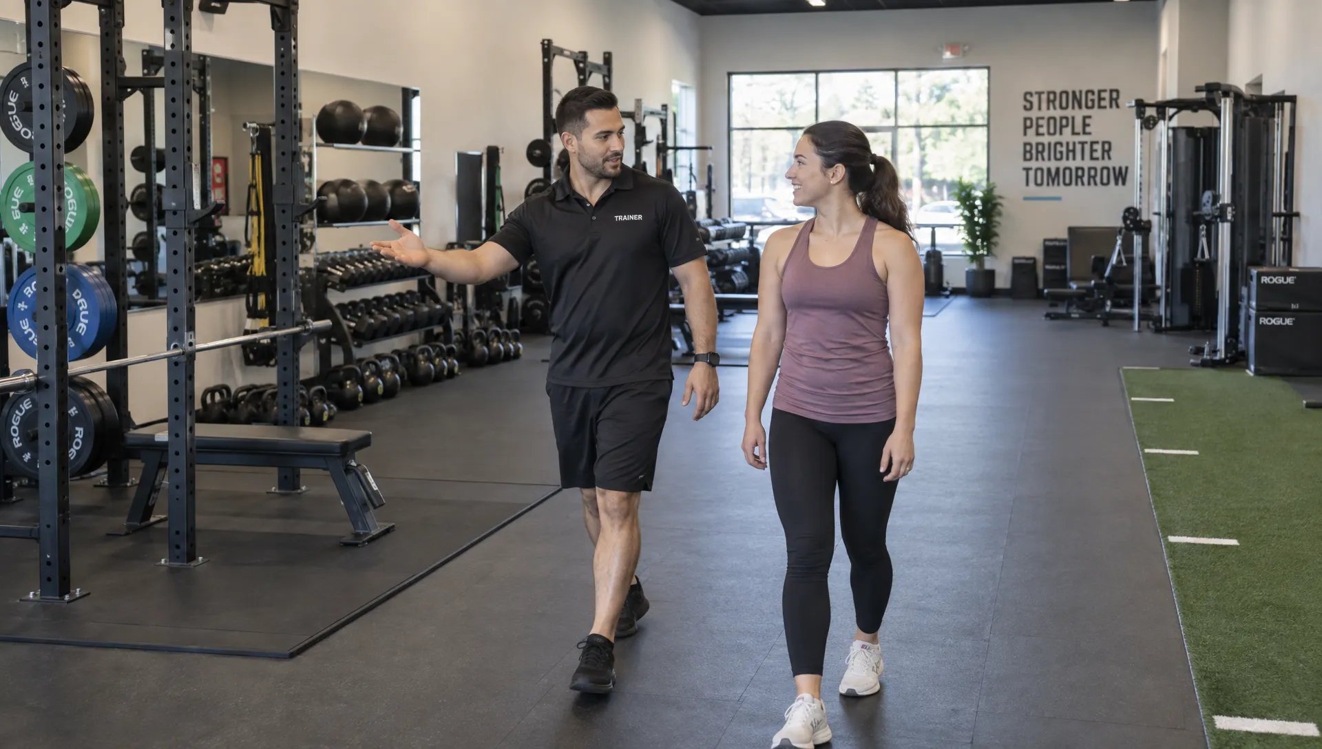 A coach shows a prospective client organized strength equipment and clear walkways during a tour of a personal training facility.
