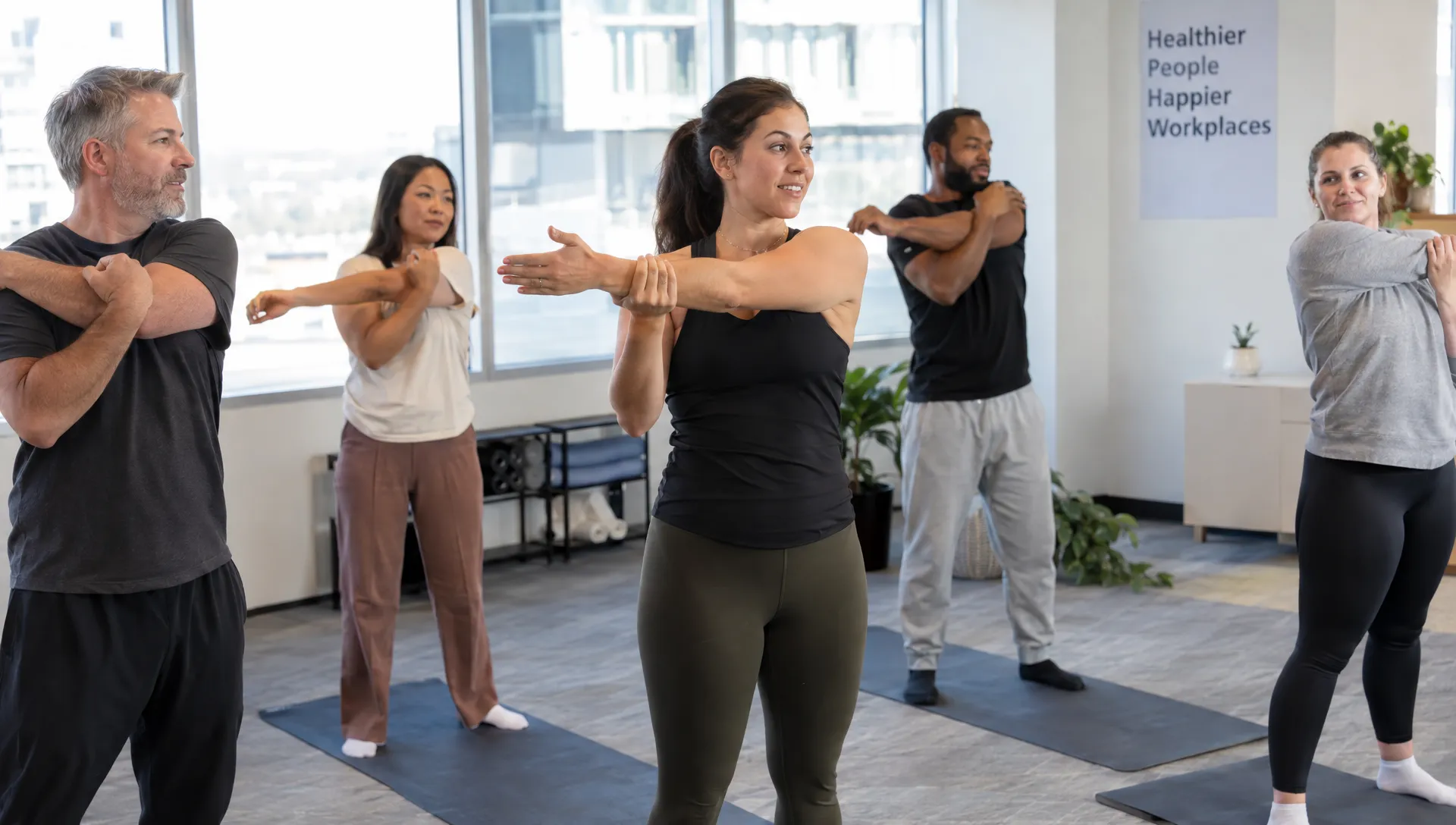 A class instructor guides adults through gentle standing mobility movements beside exercise mats during a midday work break.