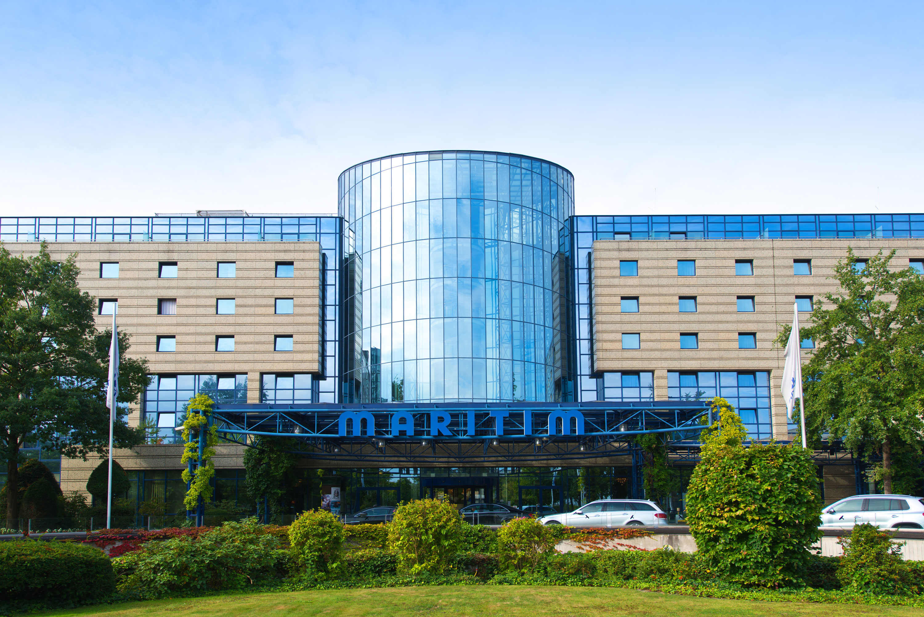 Außenansicht des Maritim Hotels mit Glasfassade und Eingangsbereich, umgeben von Bäumen und parkenden Autos.