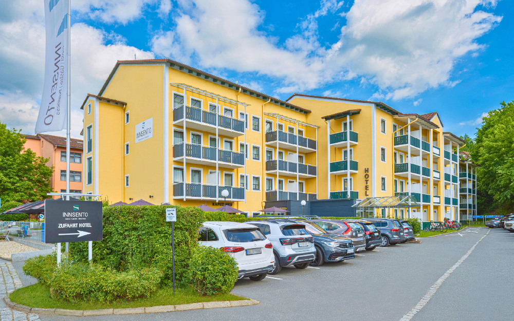 Gelbes Hotelgebäude mit Balkonen und angrenzendem Parkplatz mit mehreren geparkten Autos unter blauem Himmel.