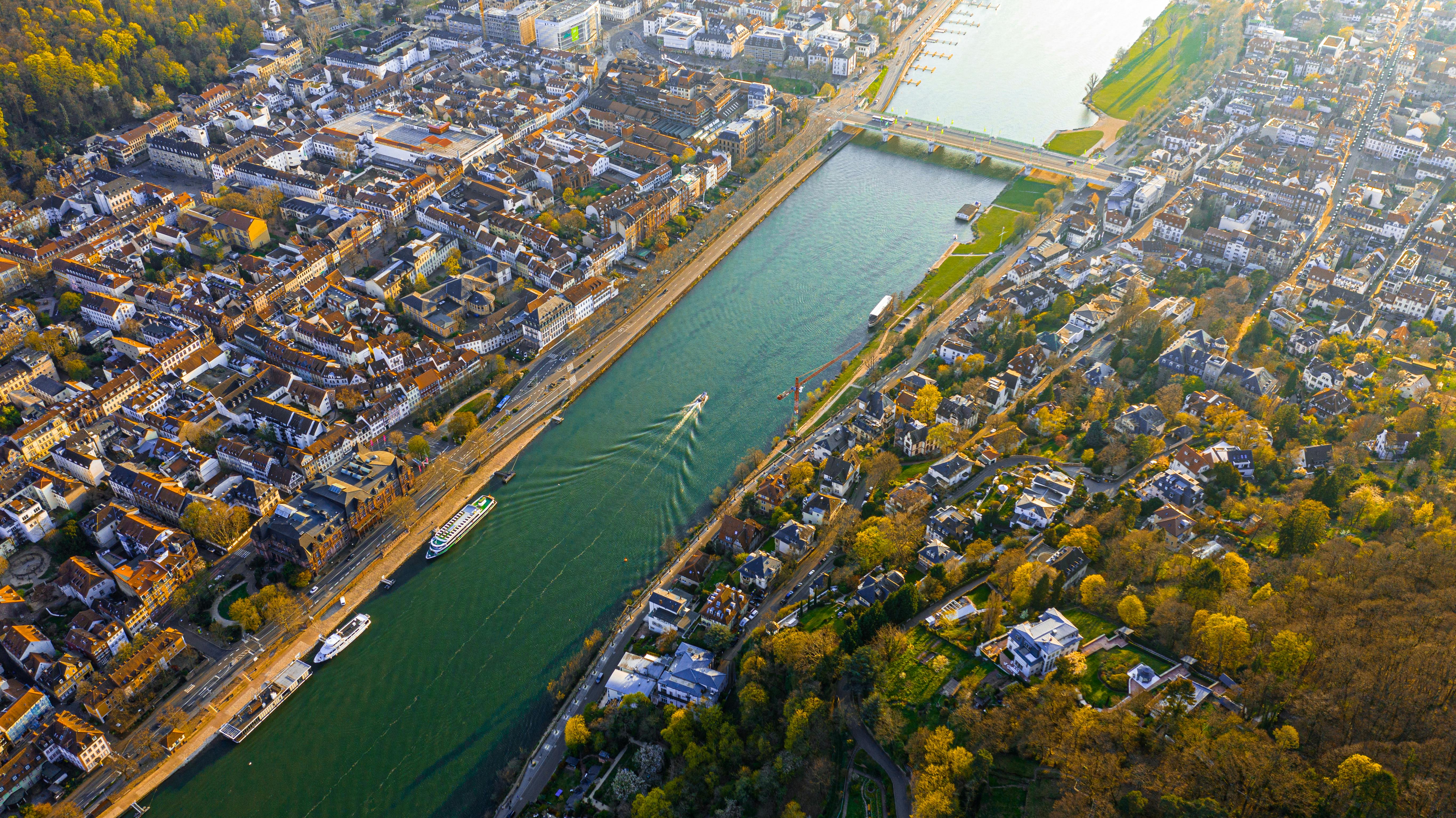 Luftaufnahme einer Stadt mit Fluss, Brücke, mehreren Booten und herbstlich gefärbten Bäumen.