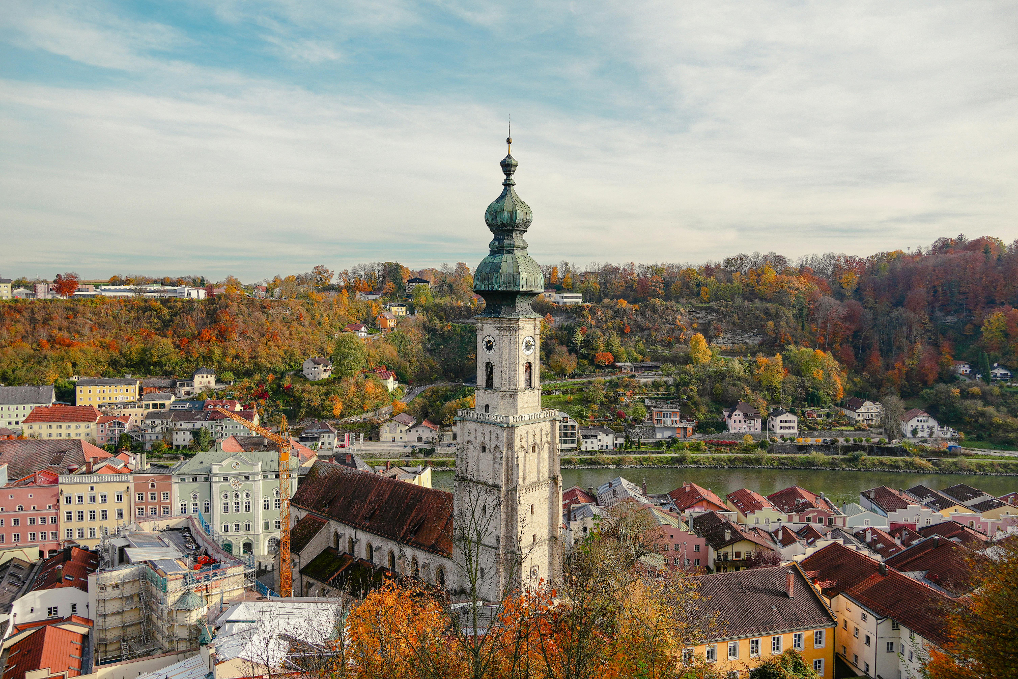 Blick auf historische Kirche mit grünem Turm in einer Stadt, umgeben von herbstlichen Bäumen und einem Fluss im Hintergrund.