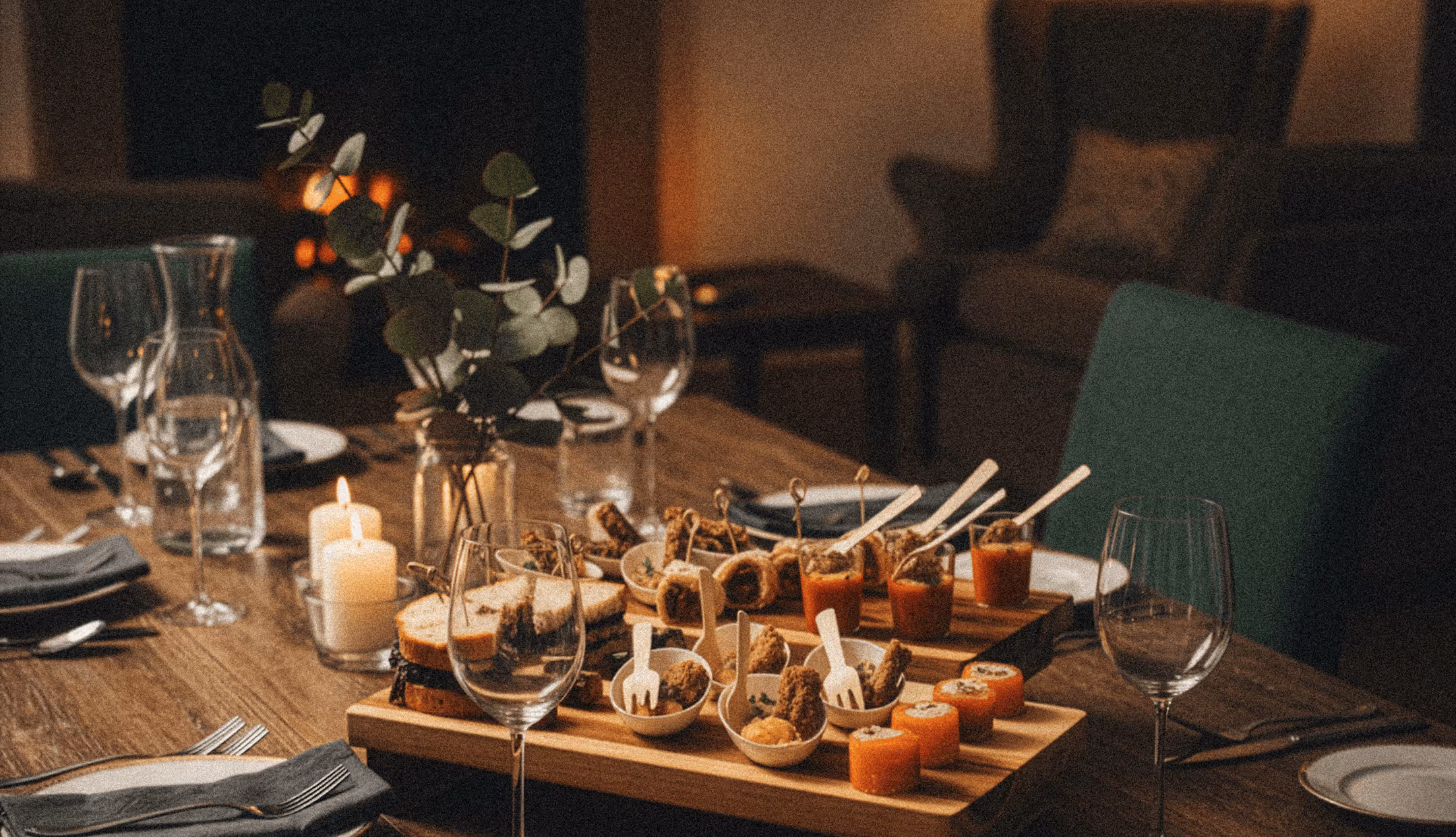 Table élégamment dressée avec des amuse-bouches variés, des verres à vin, des bougies allumées et un vase avec des branches de feuillage.