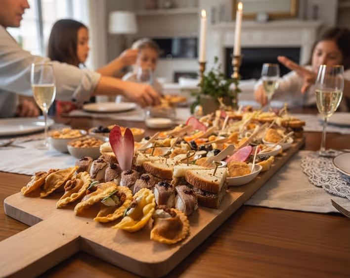 Plateau de traiteur avec divers amuse-bouches disposés sur une planche en bois, des enfants dégustant en arrière-plan autour d'une table dressée.
