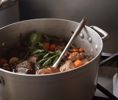Grande casserole contenant un bouillon avec morceaux de viande, carottes, branches de céleri et herbes.