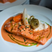 Filet de saumon avec une sauce crémeuse orange, accompagné d'asperges blanches, d'une petite carotte, et d'un légume vert, servi dans une assiette blanche avec une décoration de sauce verte.