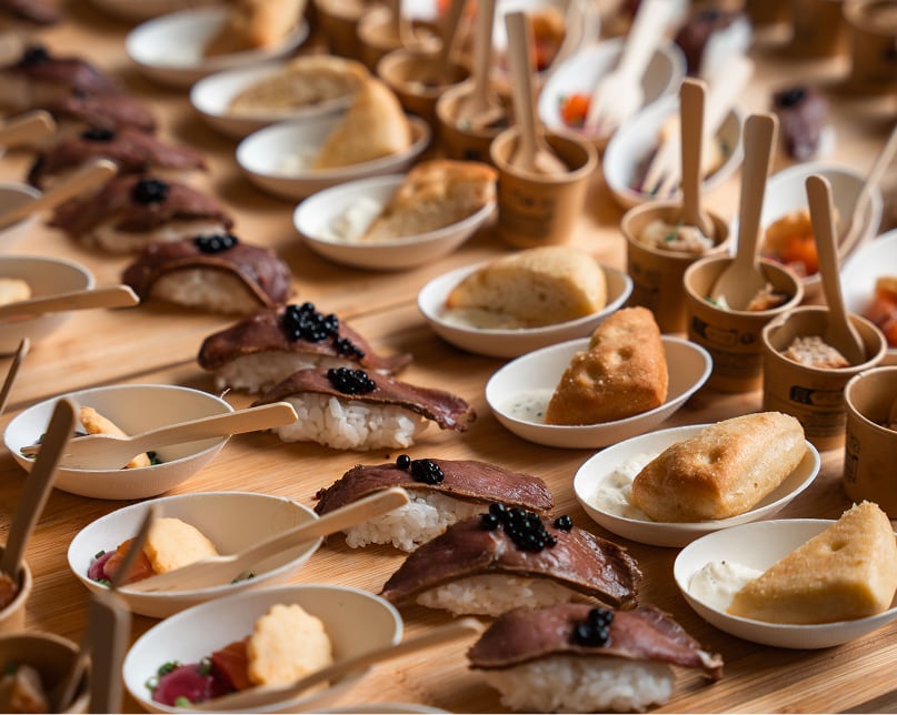 Plateaux de buffet traiteur avec sushis garnis de viande et perles noires, petits pains frits avec sauce blanche, et diverses petites portions dans des bols et gobelets, présentés sur une table en bois.
