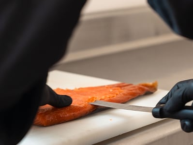 Personne portant des gants noirs découpe un filet de saumon frais sur une planche blanche avec un couteau tranchant.