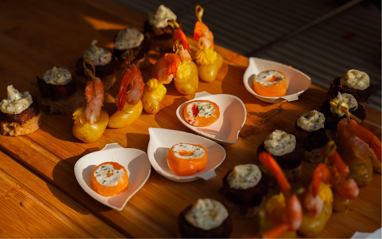 Assortiment de bouchées apéritives sur plateau en bois, comprenant des crevettes sur pommes de terre, des roulés de saumon au fromage frais, et des morceaux de pain avec une crème aux herbes.