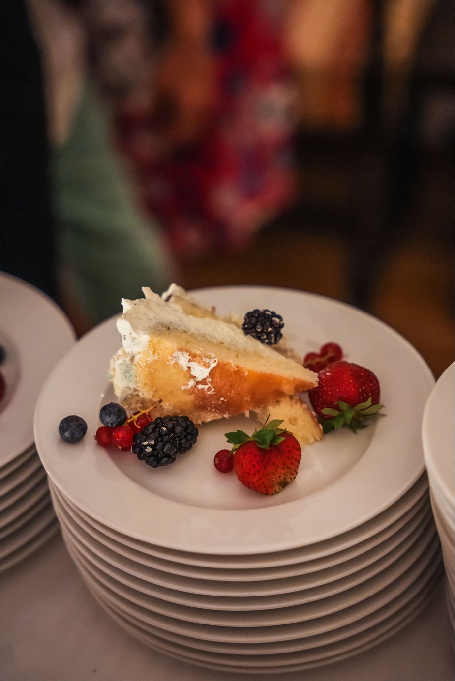 Une part de gâteau à la crème blanche garnie de fraises, mûres, myrtilles et groseilles rouges, posée sur une assiette blanche sur une pile d'assiettes similaires.